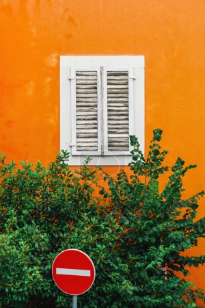 Téléchargez les photos : Scène urbaine colorée - Pas de panneau de signalisation à côté d'un vieux bâtiment orange avec fenêtre blanche fermée et cime des arbres verte, mise au point sélective - en image libre de droit