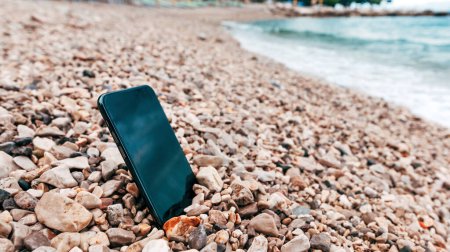 Foto de Nuevo teléfono móvil moderno perdido en la playa junto al mar en guijarros, enfoque selectivo - Imagen libre de derechos