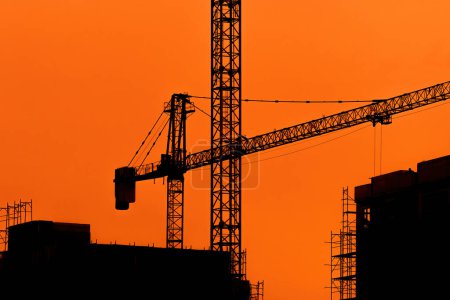 Foto de Silueta de construcción grúa, andamios y edificios en puesta de sol contra cielo anaranjado. Espacio de copia incluido. - Imagen libre de derechos