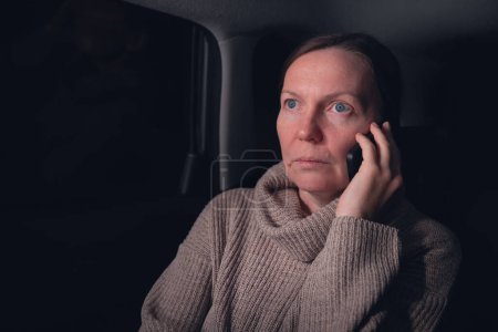 Photo for Woman talking on mobile phone from the back seat of the car while on a road trip at night, selective focus - Royalty Free Image