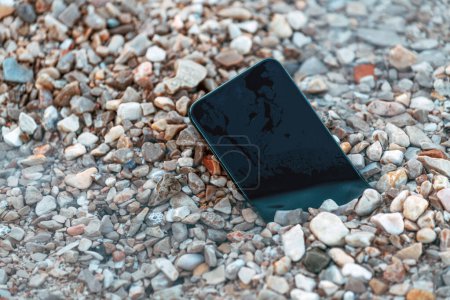Téléchargez les photos : Appareil de téléphone intelligent perdu dans les galets sur la plage de bord de mer le matin d'été, mise au point sélective - en image libre de droit