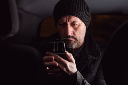 Téléchargez les photos : Adulte caucasien mâle lecture texte message sur téléphone cellulaire tandis que assis sur le siège arrière de la voiture la nuit, mise au point sélective - en image libre de droit