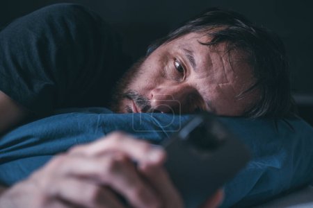 Mann mit Smartphone spät in der Nacht im Bett liegend, Schlaflosigkeit, selektiver Fokus