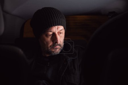 Photo for Serious worried man thinking at back seat of the car at night. Adult male wearing black cap and jacket and thinking. Selective focus. - Royalty Free Image