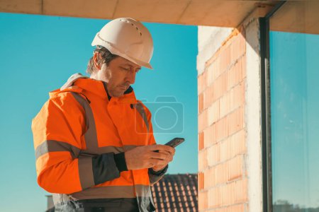 Téléchargez les photos : Ingénieur en construction utilisant le téléphone intelligent mobile sur place pendant l'inspection, foyer sélectif - en image libre de droit