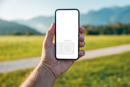 Téléchargez les photos : Modélisation de smartphone, homme tenant un téléphone portable avec écran blanc vierge comme espace de copie sur un paysage de prairie alpine tranquille le matin d'été, mise au point sélective - en image libre de droit
