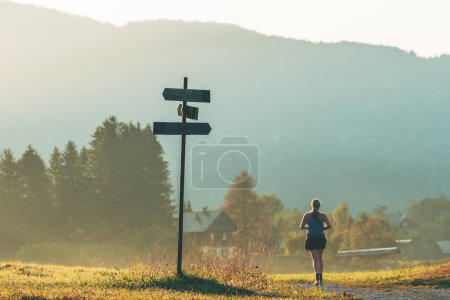 Téléchargez les photos : Routine de jogging du matin, joggeuse méconnaissable qui court sur un sentier de gravier à travers un magnifique paysage de campagne alpine le matin d'été, accent sélectif - en image libre de droit