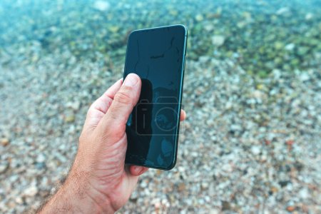 Téléchargez les photos : Homme tenant humide téléphone portable étanche trouvé dans l'eau de mer, mise au point sélective - en image libre de droit