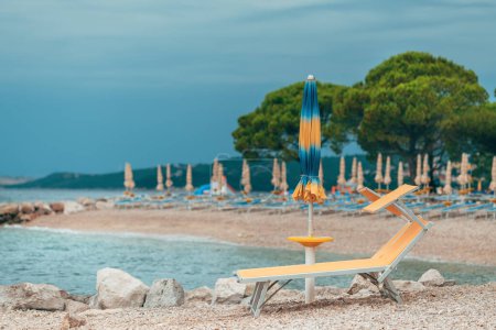 Folded beach umbrellas and empty deck chairs on town beach in Crikvenica, Croatia. Overcast weather and cloudy days during poor touristic season, selective focus