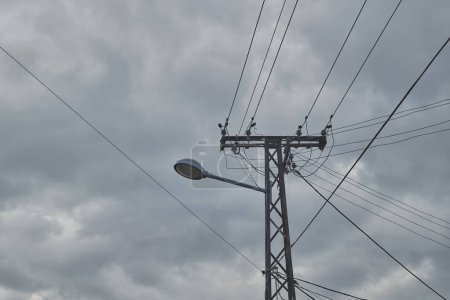 Street light mounted on a metal electrical post, accentuated by intertwining power lines against overcast sky. Selective focus.