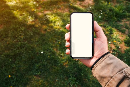 Téléchargez les photos : Modèle de téléphone intelligent, main masculine tenant téléphone portable avec écran blanc vierge dans le parc le matin du printemps, mise au point sélective - en image libre de droit