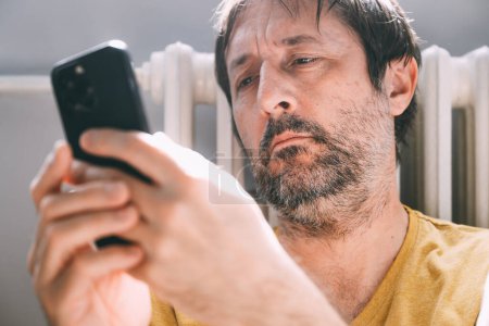 Téléchargez les photos : Homme utilisant un téléphone portable à la maison tout en s'appuyant sur le radiateur de chauffage, foyer sélectif - en image libre de droit