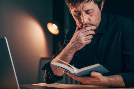 Téléchargez les photos : Homme d'affaires lisant un livre assis au bureau tard le soir, objectif sélectif - en image libre de droit