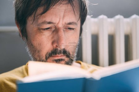 Téléchargez les photos : Homme caucasien adulte avec barbe lecture livre relié bleu dans le salon à la maison le matin, mise au point sélective - en image libre de droit