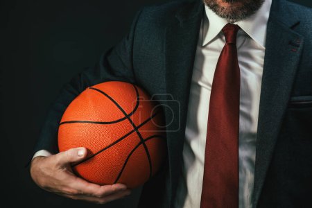 Téléchargez les photos : Coach de basket-ball en costume d'affaires élégant tenant la balle sur fond sombre, mise au point sélective - en image libre de droit
