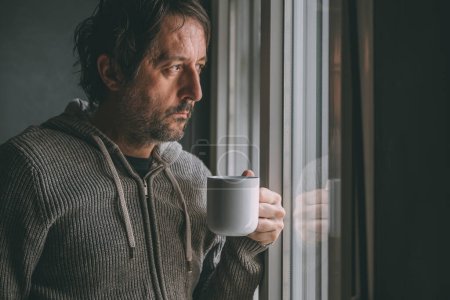 Photo for Pensive adult male drinking coffee in the morning by the apartment window while looking out and thinking. Caffeine dose for starting of the day. Selective focus. - Royalty Free Image