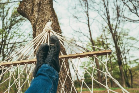Téléchargez les photos : Homme allongé et relaxant dans un hamac de jardin attaché à un arbre, portant un jean et des baskets noires sales, foyer sélectif - en image libre de droit