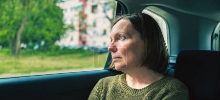 Photo for Portrait of serious mature adult female thinking at the back seat of a car while driving through residential neighborhood, selective focus - Royalty Free Image