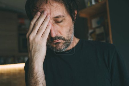 Foto de Hombre adulto deprimido con dolor de cabeza masajeando la frente en la cocina casera, enfoque selectivo - Imagen libre de derechos