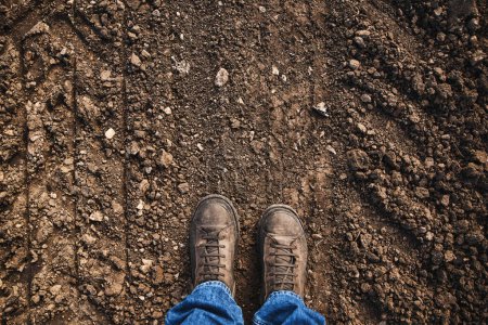 Téléchargez les photos : Vue de dessus des pieds de fermier en bottes debout sur le chemin de terre de campagne, espace de copie inclus - en image libre de droit