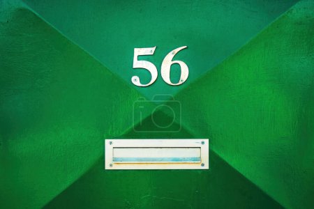 House number 56 on green metal door and mailbox slot. Fifty six in worn metallic numerals. Copy space included.