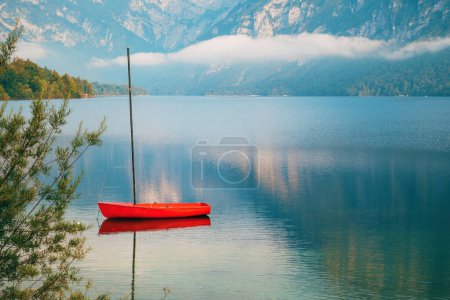 Red boat floating on tranquil lake Bohinj surface in summer morning, selective focus