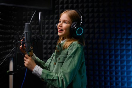 Téléchargez les photos : Jeune adolescente chanteuse star portant des écouteurs chantant une nouvelle chanson avec microphone au studio de disques - en image libre de droit