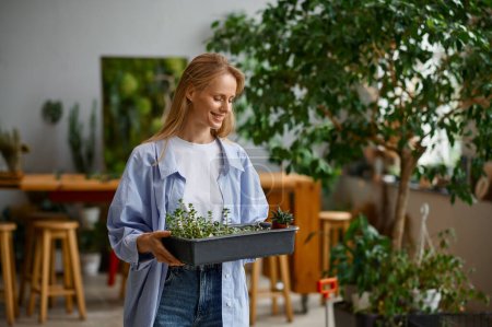 Photo for Young female florist worker enjoying her job. Portrait of smiling woman walking with plants in plastic box. Floristic workshop concept - Royalty Free Image