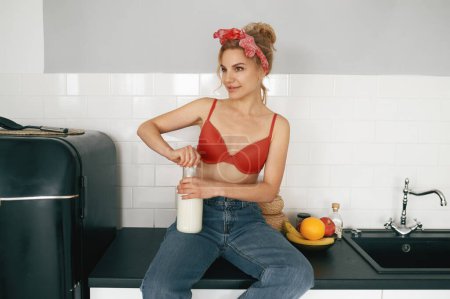 Téléchargez les photos : Jeune femme en lingerie ouvre bouteille avec du lait et souriant tout en étant assis sur la table de cuisine - en image libre de droit