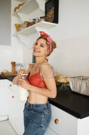 Foto de Mujer joven en lencería abre una botella con leche y sonríe en la cocina - Imagen libre de derechos