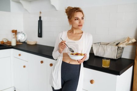 Téléchargez les photos : Jeune femme rêveuse mangeant des céréales avec du lait petit déjeuner sain sur la cuisine à la maison après l'entraînement de fitness matinal - en image libre de droit