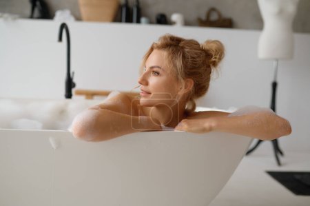 Téléchargez les photos : Portrait d'une jeune femme songeuse prenant son bain et détournant les yeux. Belle femelle relaxante tout en étant allongée dans la baignoire. Procédure de spa à la maison et hygiène - en image libre de droit