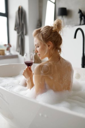 Photo for Sensual relaxed woman drinking red wine while taking bath with foam rear view Home spa and rest on weekend or calm evening for stress relief - Royalty Free Image