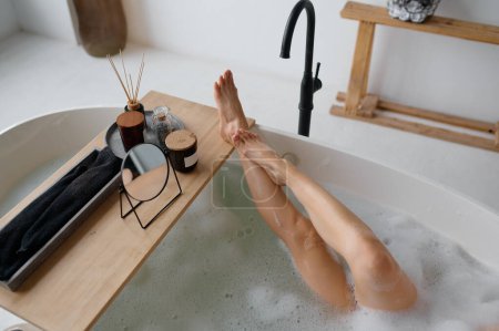 Téléchargez les photos : Vue d'en haut sur patte femelle mince humide dans un bain mousseux. Accueil spa de jour, procédure d'arôme dans la baignoire, se reposer et se rafraîchir après le travail, moment pour soi-même - en image libre de droit