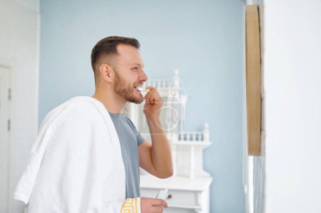 Téléchargez les photos : Jeune homme souriant avec brosse à dents nettoyer les dents et miroir de regard dans la salle de bain à la maison. routine d'hygiène quotidienne - en image libre de droit