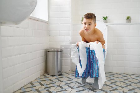 Téléchargez les photos : Petit garçon fronçant les sourcils tenant du papier assis sur les toilettes et essayant de faire caca. Constipation infantile, problèmes digestifs - en image libre de droit