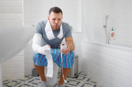 Téléchargez les photos : Froncement des sourcils jeune homme adulte assis sur les toilettes souffre de constipation tenant du papier blanc à la main pour révéler la douleur. Guy vêtu de vêtements domestiques pose dans la salle de repos - en image libre de droit
