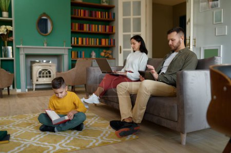 Photo for Happy family spending time at home together in evening. Father and mother using digital gadget, little son reading book sitting on floor - Royalty Free Image