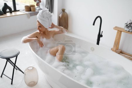 Foto de Retrato de mujer hermosa con toalla en la cabeza tomando baño de aroma caliente con burbujas de espuma. Descanse y relájese en casa - Imagen libre de derechos