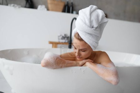 Téléchargez les photos : Portrait de femme magnifique portant une serviette sur la tête prenant un bain d'arôme chaud avec des bulles de mousse. Repos et détente à la maison - en image libre de droit