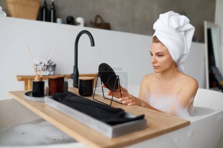Foto de Joven hermosa mujer con toalla de baño spa disfruta del procedimiento de cuidado de la piel de la cara mientras está sentado en una bañera caliente con burbujas de espuma en casa - Imagen libre de derechos