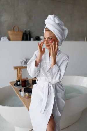 Foto de Mujer joven en toalla aplicando parche cosmético de belleza debajo de los ojos para eliminar las arrugas después de bañarse o ducharse. Cuidado de la piel, tratamiento de belleza, cosmetología, concepto de dermatología - Imagen libre de derechos