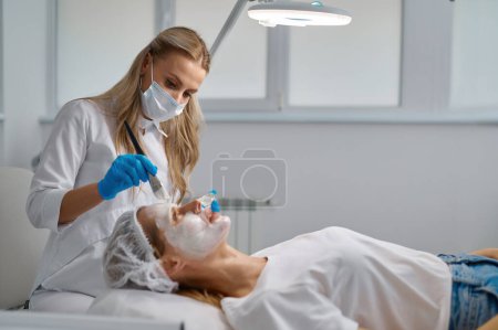 Foto de Esteticista aplicación de mascarilla de arcilla en la cara de mujer vista lateral. Concepto de tratamiento de rejuvenecimiento - Imagen libre de derechos