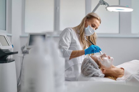 Téléchargez les photos : Beautician applying clay face mask on woman face side view. Rejuvenation treatment - en image libre de droit
