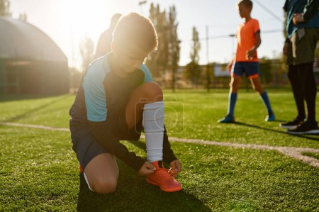 Foto de Closeup boy little football player tying shoelaces of training boots sitting on green grass soccer field before match - Imagen libre de derechos