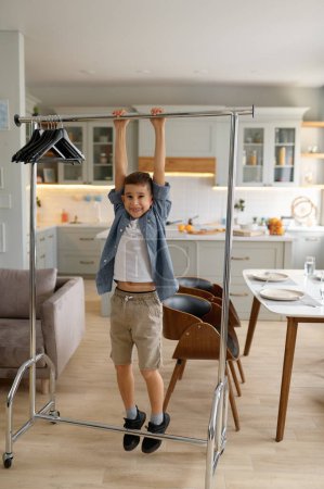 Foto de Little preteen boy child happily smiling while hanging on clothes hanger. Cozy home interior and naughty mischievous kid in family - Imagen libre de derechos