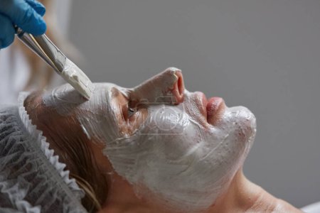 Téléchargez les photos : Closeup side view on cosmetologist brush applying clay mask on female client face - en image libre de droit