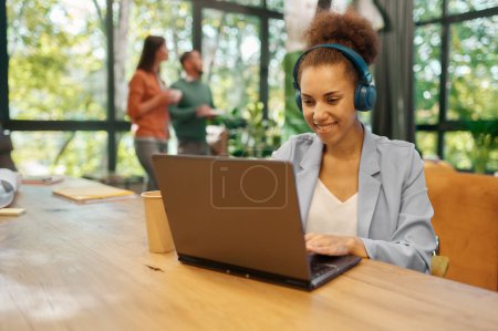 Photo for Coworking space concept. Focus on young business woman freelancer wearing headphones using laptop computer for online work and video conferencing at shared open workplace - Royalty Free Image
