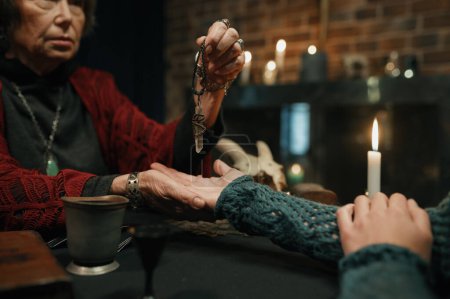 Téléchargez les photos : Mature witch using amethyst crystal pendulum to predict future for young female client. Palmistry, fortune telling and occultism concept - en image libre de droit