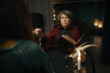 Téléchargez les photos : Gypsy forecaster reading spell for client using witchcraft book and magic pendulum psychic tool selective focus - en image libre de droit